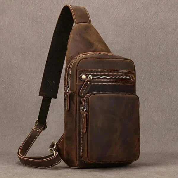Brown leather crossbody bag with carrying strap, zipper opening, two external pockets, gray background.