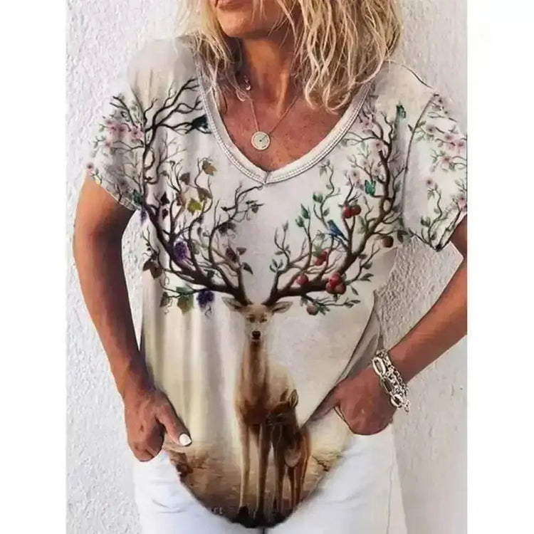 Woman wearing a short sleeve T-shirt with an intricate deer and floral graphic, hands in pockets, against a white wall.