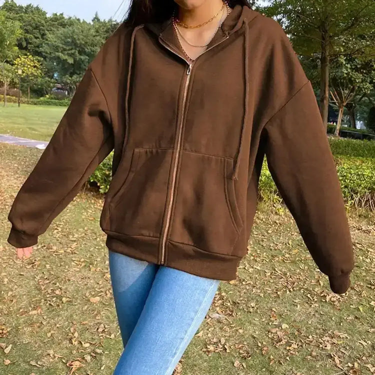 A woman wearing a brown long-sleeved casual hoodie jacket with an oversized hood and adjustable collar is standing outdoors in a park.