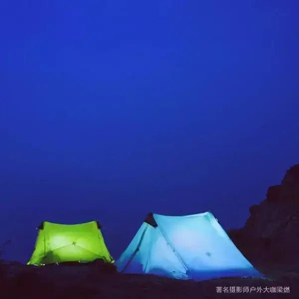 Two ultralight camping tents glowing in the dark under a clear blue night sky on rocky terrain.