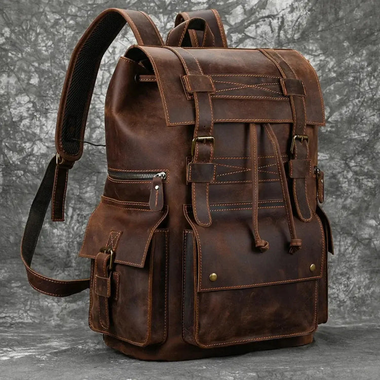 A brown leather backpack with a flap top secured by buckle straps and a drawstring, featuring various external pockets and adjustable shoulder straps, against a mottled gray backdrop.