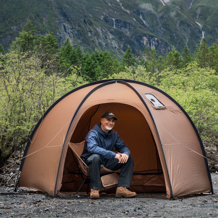 Man sitting inside a brown tent with mountains and greenery in the background