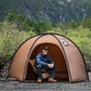 Man sitting inside a brown tent with mountains and greenery in the background