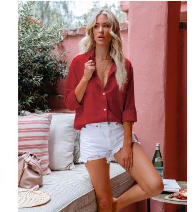 woman in red linen tunics sitting on a couch, wearing white shorts, with summer decor and greenery in the background.