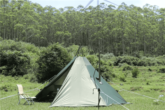 Pyramidal green camping tent set up on a green slope, surrounded by trees and wind turbines in the background