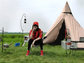 A spacious, pyramid-shaped tent, pitched on a grassy field with a woman sitting in front.